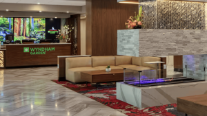 The hotel lobby features a sleek, contemporary design centered around a striking stone fireplace. A beige sectional sofa provides ample seating space, accentuated by a vibrant red-patterned rug that enlivens the floor. A sturdy wooden coffee table adds practicality and style to the setting. The reception area displays the Wyndham Garden sign prominently, merging seamlessly with the refined backdrop of Redwood Valley’s charm. This welcoming atmosphere is perfect for guests checking in with their furry companions as they plan their stay in comfort and style.